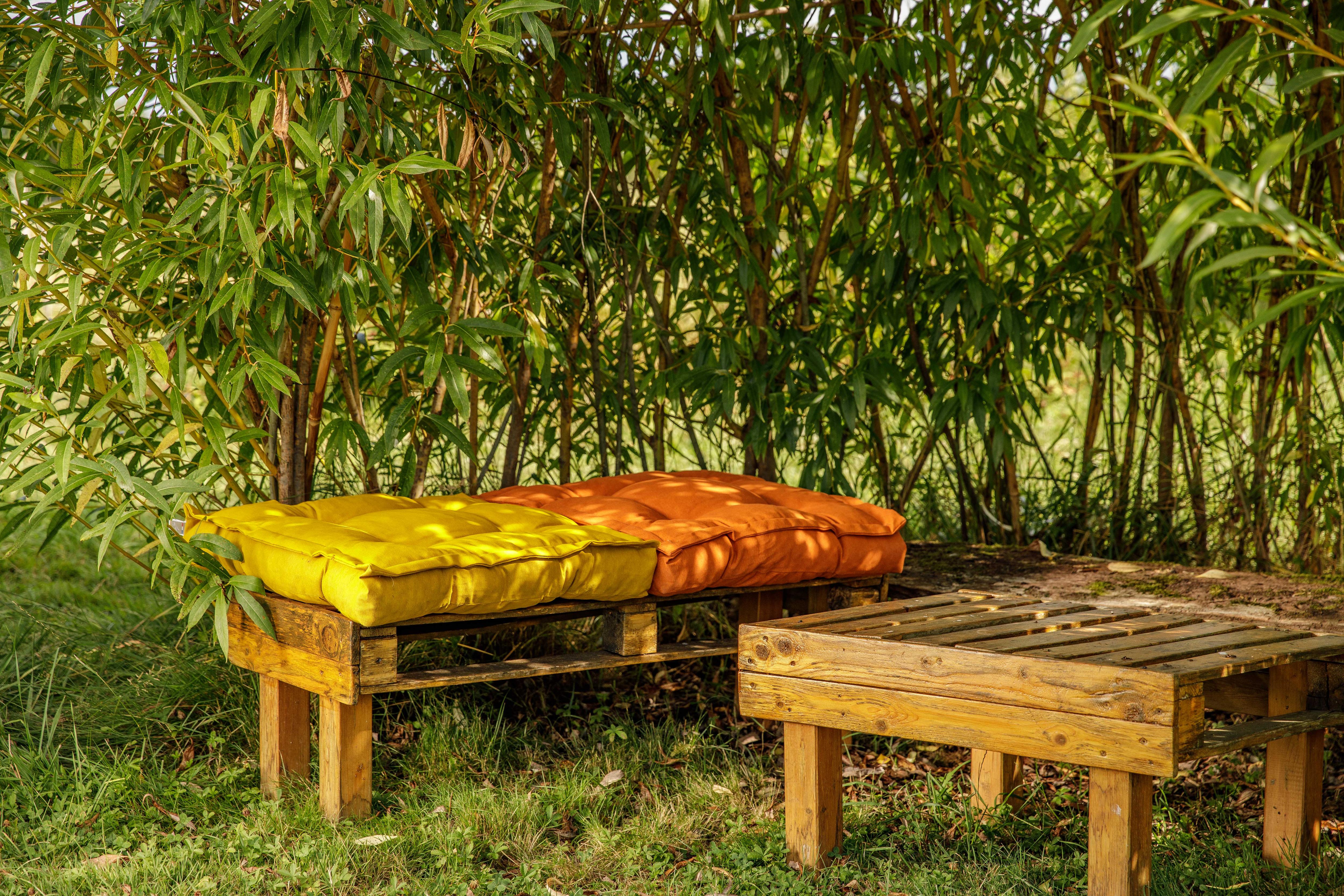 Un petit salon dans un jardin avec des coussins jaune et orange sous un arbres d'ombrage