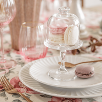 Une table dressée avec des assiettes blanches et un plat à pâtisserie garni de macarons sous cloche