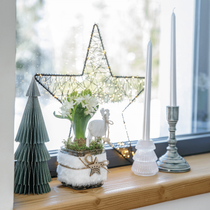 Un rebord de fenêtre en hiver avec une composition de jacinthes, une déco sapin en papier, une étoile lumineuse et des bougies cierges