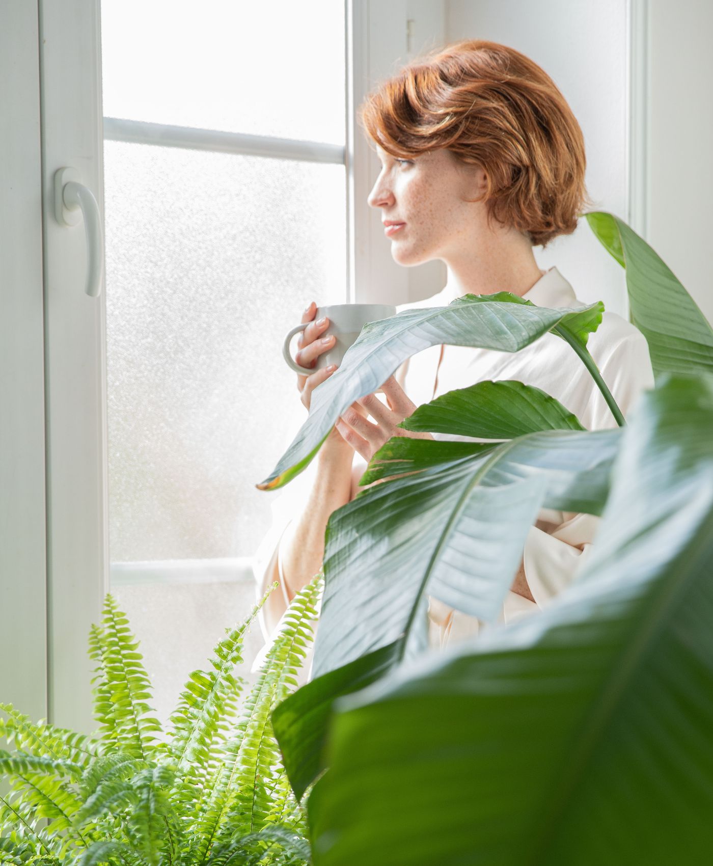 Une femme qui regarde par la fenêtre avec un café à la main et des plantes autour d'elle