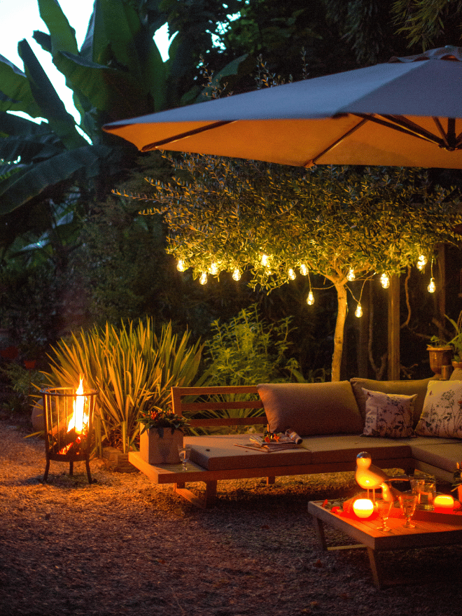 Une terrasse extérieure aménagée avec un brasero, un parasol, un salon de jardin et des guirlandes