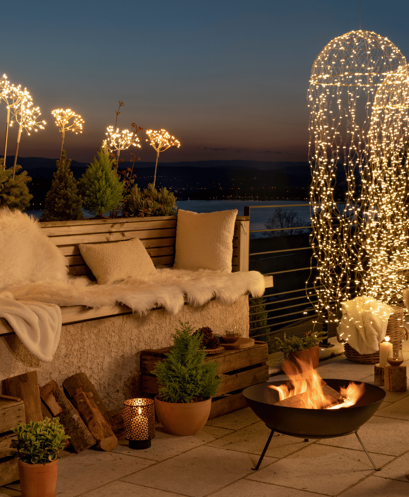 Une terrasse de nuit aménagée avec un brasero, des coussins, des plaids, des luminaires et des végétaux