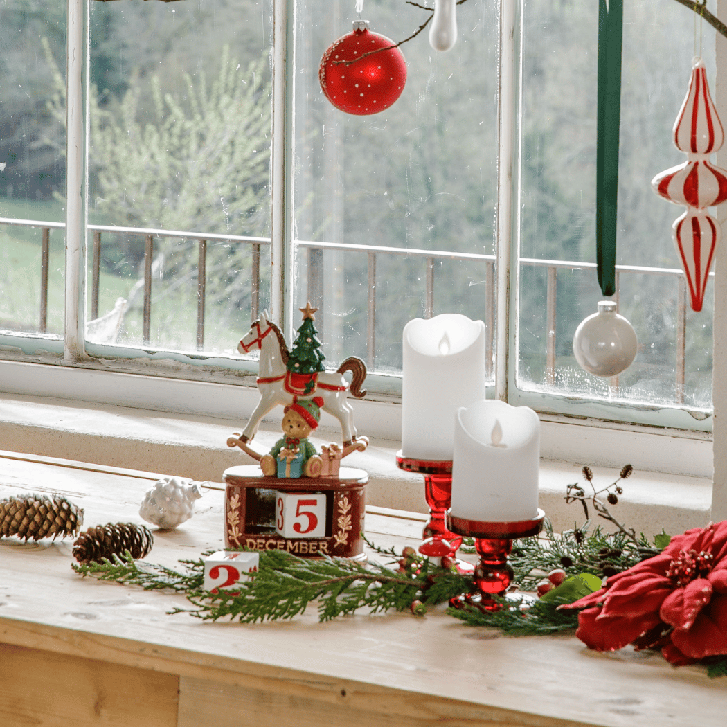 Sur un meuble devant une fenêtre, il y a un calendrier de l'avent décoratif avec un cheval à bascule et des décorations de couleur rouge et blanches