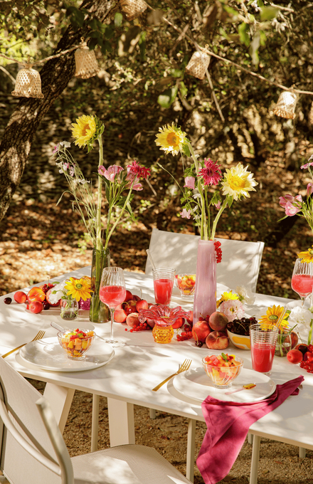 Une table d'été sous un arbre avec une guirlande solaire suspendue, des fleurs dans des vases et des fruits dans les assiettes