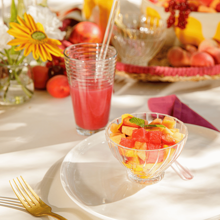 Des fruits d'été, pastèques et melons, coupés dans une coupe posée dans une assiette blanche, accompagnés d'un jus de fruits coloré dans un verre