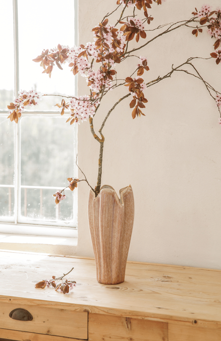 Un grand vase rose en céramique posée sur un meuble en bois près d'une fenêtre avec une branche de cerisier en fleurs