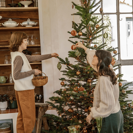 Un ado sur un tabouret avec un panier à la main, et sa maman en train de mettre des décorations de Noël sur le sapin