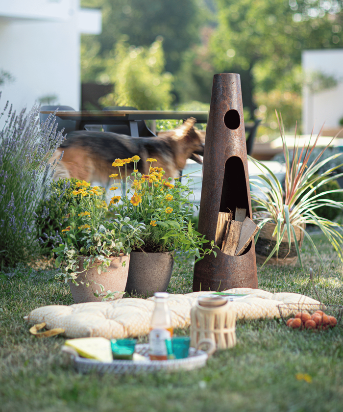 Un brasero décoratif dans le jardin avec des coussins, un plateau, des boissons et des végétaux